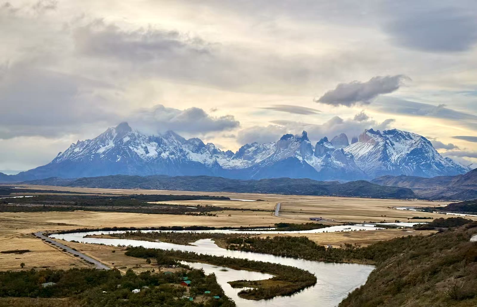 这整片绵延壮阔的山脉被总称为百内群峰(Cordillera Paine 或 Paine Massif)。它是智利百内国家公园最核心的标志性景观。• 画面左侧： 那座体量最为庞大、顶部覆盖着宽阔厚重冰雪的山体，是群峰的最高点大百内峰(Paine Grande)。• 画面中右侧： 那些连绵起伏、如刀劈斧削般尖锐，且带有黑白双色岩层特征的山峰，正是标志性的百内角(Cuernos del Paine)。还有那被云雾环绕的像手指尖的著名三塔中的中塔。